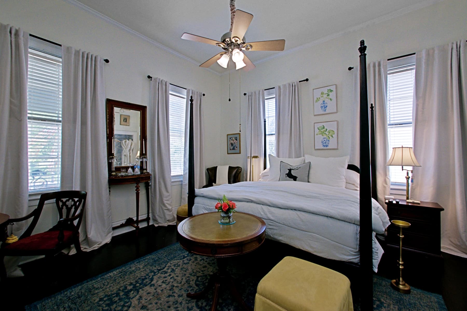 An elegant bedroom featuring a dark wood four-poster bed with crisp white linens and a grey throw blanket. In the foreground, two light blue chairs face a round wooden table topped with a small vase of red flowers, while a dark leather wingback chair with a yellow ottoman sits to the left. The room is accented with long white curtains framing the windows and several framed botanical prints on the walls.
