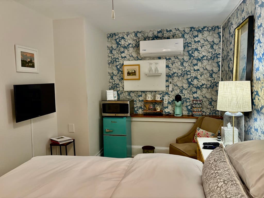 A small kitchenette area located within a bedroom, featuring a vibrant teal mini-fridge with a microwave sitting on top. The back wall is covered in blue and white floral wallpaper and holds a mounted air conditioning unit, a small white shelf with glassware, and coffee-making supplies. A wall-mounted television is visible on the adjacent plain white wall.