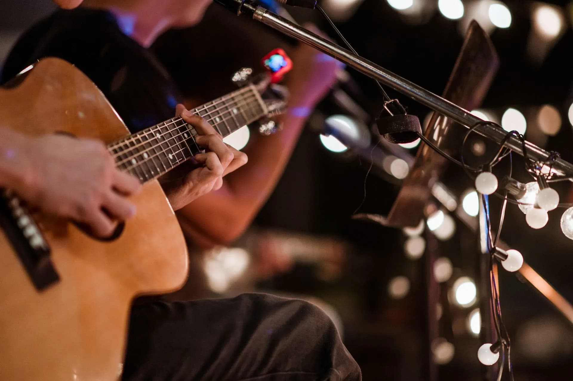 A close-up of a musician's hands playing an acoustic guitar, with blurred lights in the background.