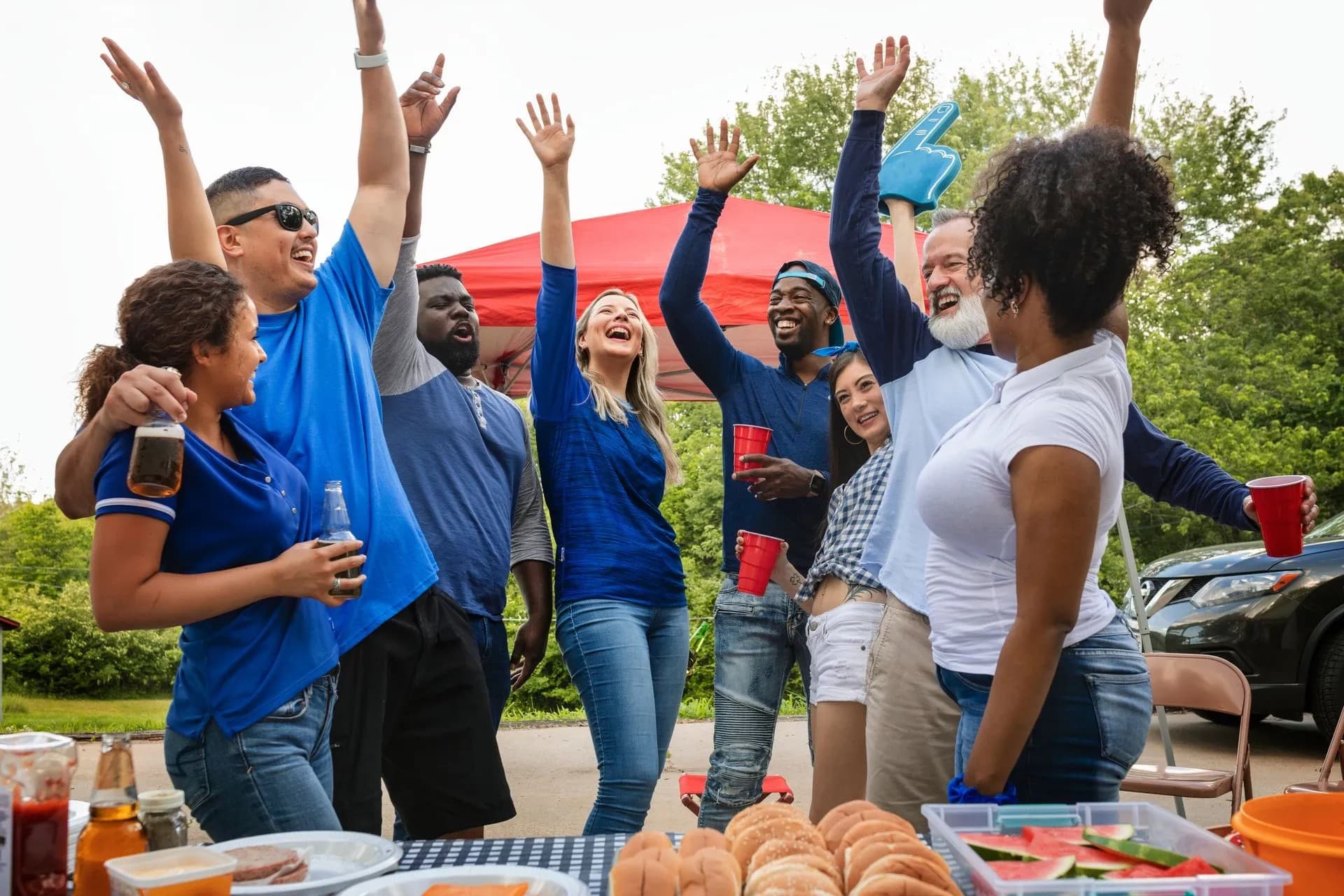 A group of diverse friends joyfully celebrate outdoors, raising their arms in excitement beside a table of food and drinks.