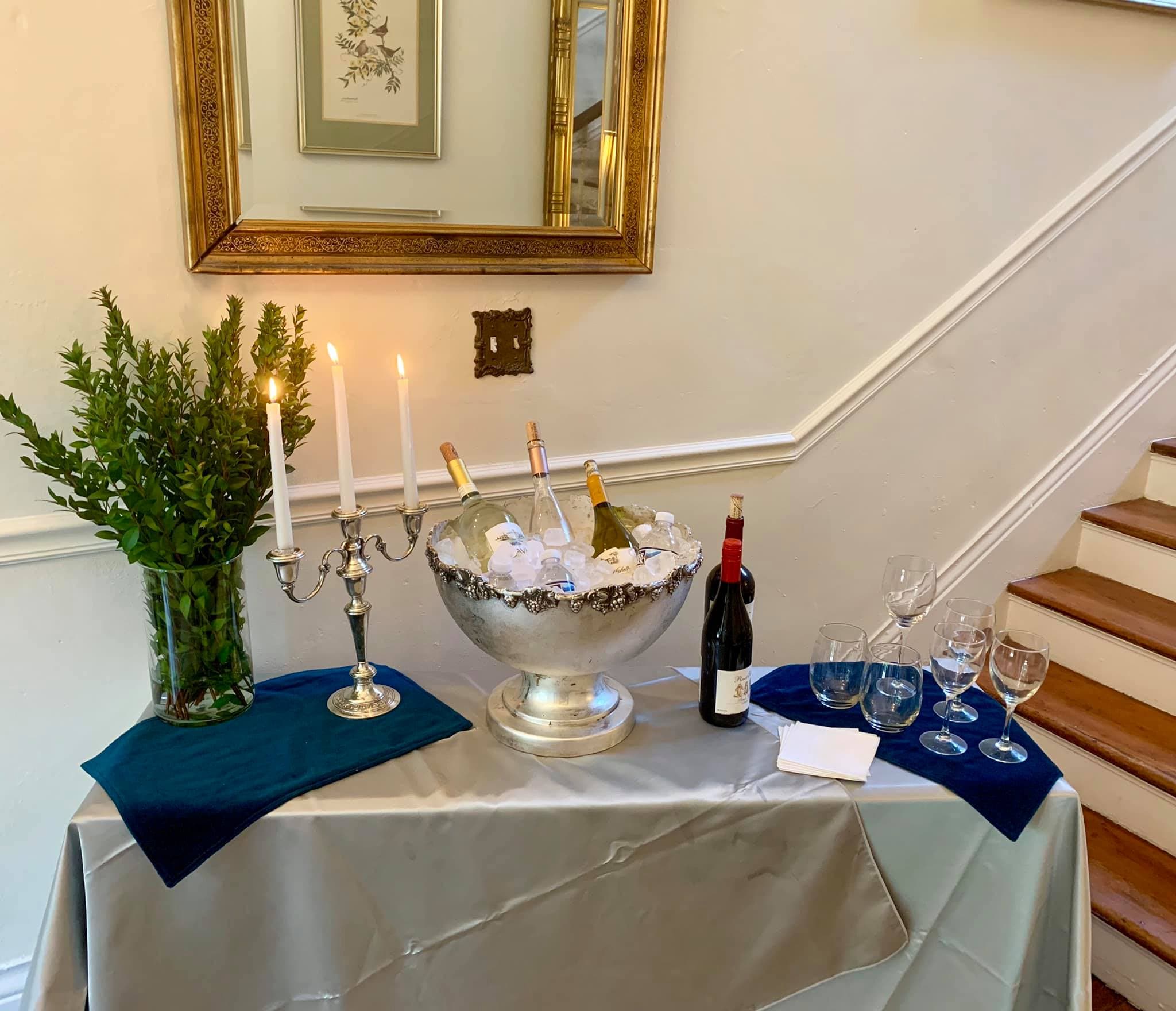 A beautifully arranged table with a silver ice bucket holding champagne bottles, a bottle of red wine, candles, glasses, and greenery.