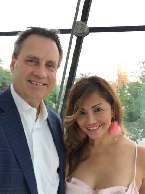 A man and a woman pose for a selfie with a cityscape in the background.