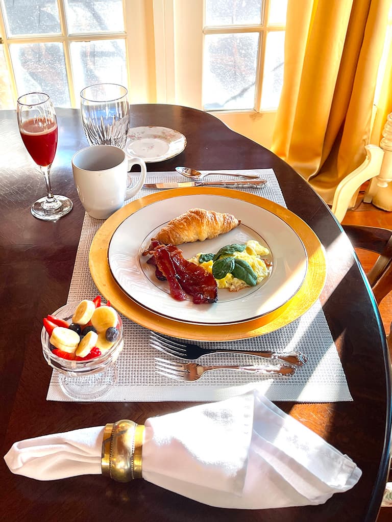A beautifully arranged breakfast plate featuring scrambled eggs, bacon, a croissant, and a fruit bowl, accompanied by beverages on a wooden table near a window.