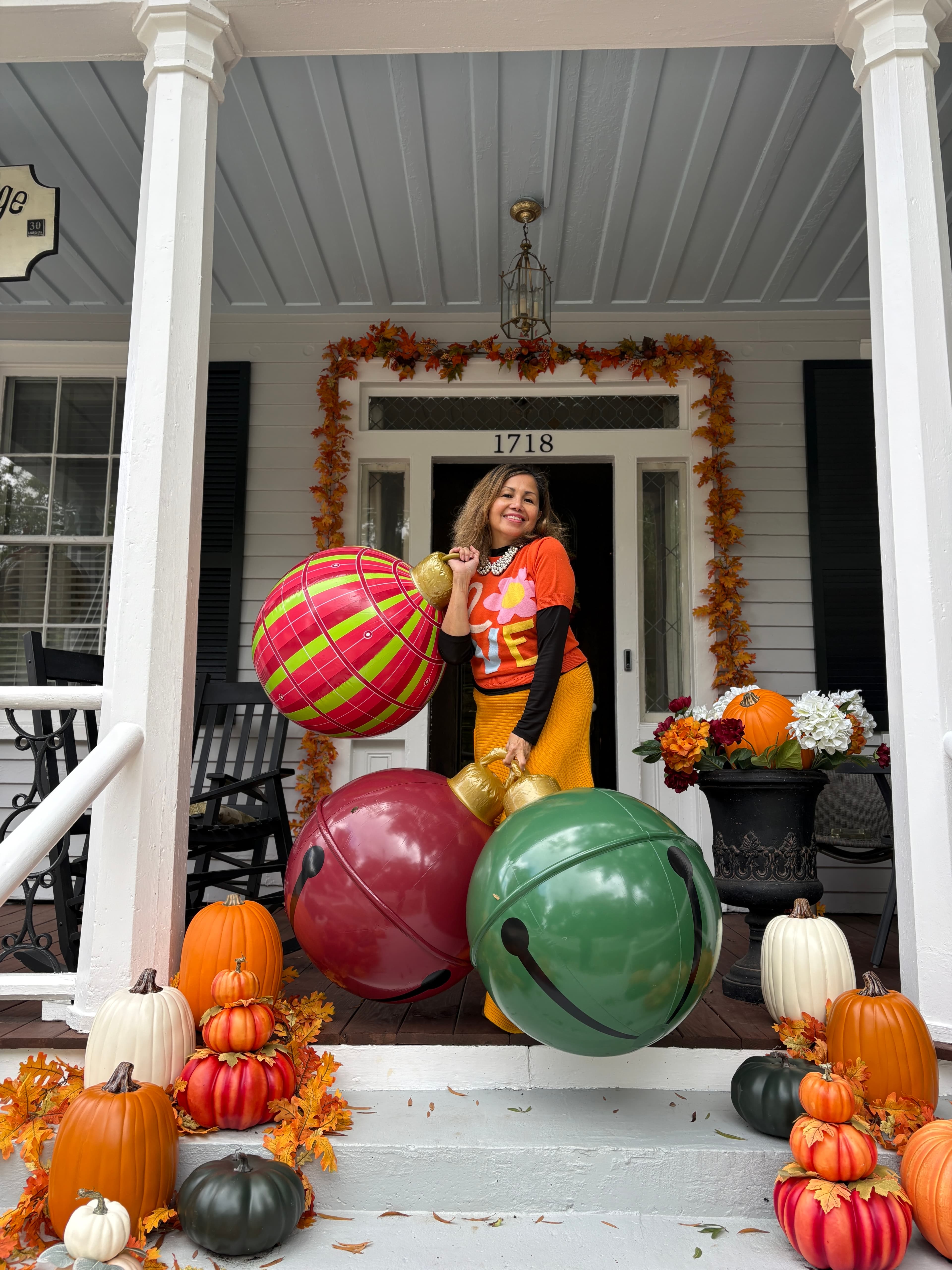 A smiling woman in colorful attire holds large decorative ornaments on a porch adorned with pumpkins and autumn foliage.