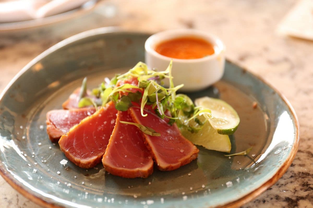 A plate of sliced seared tuna garnished with microgreens, lime, and a small bowl of dipping sauce.