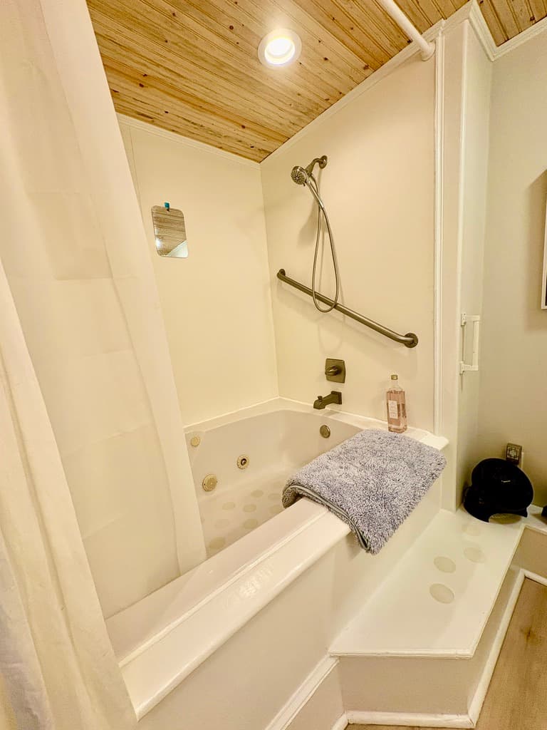 A close-up shot of a white jetted bathtub and shower combination with a wood-plank ceiling. A blue towel is draped over the edge of the tub next to a bottle of soap, and a dark metal safety grab bar is mounted on the wall near the showerhead.