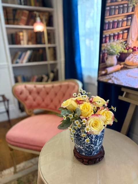 A vase of yellow roses and daisies sits on a table next to a pink upholstered chair in a cozy, well-lit room.