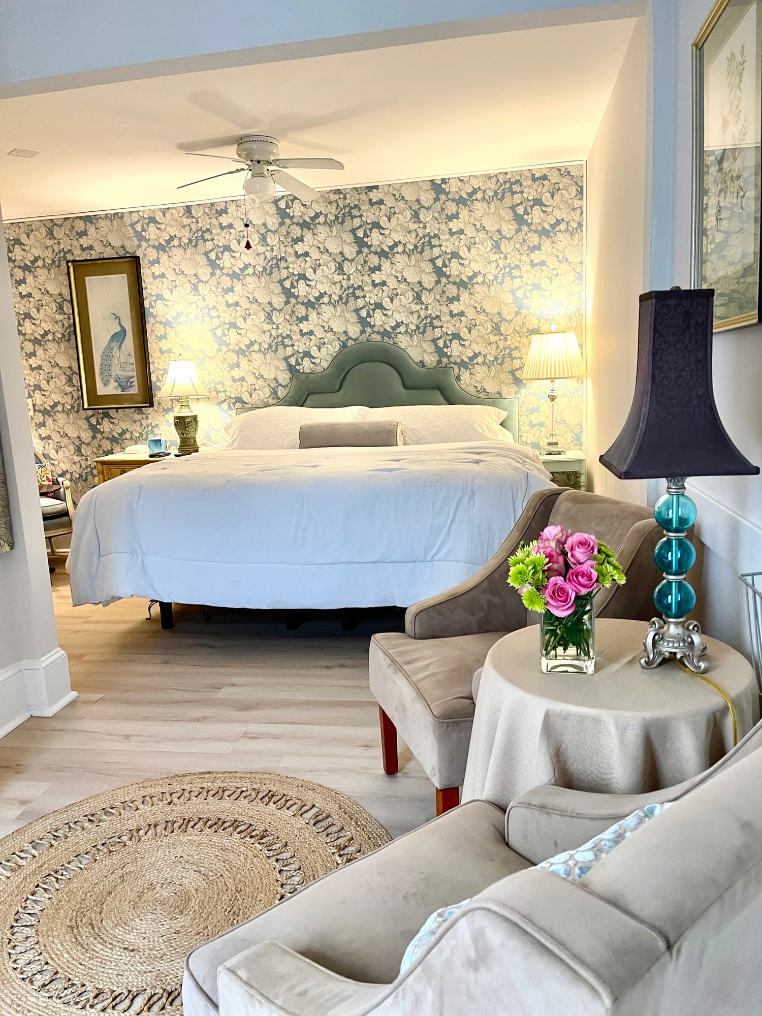 A brightly lit bedroom viewed from an adjacent sitting area. The bedroom features a bed with white linens and a pale green scalloped headboard set against a blue and white floral wallpaper accent wall. In the foreground, two beige velvet armchairs face a small round table draped in a tan cloth, which holds a vase of pink flowers and a lamp with a blue glass base and dark purple shade.