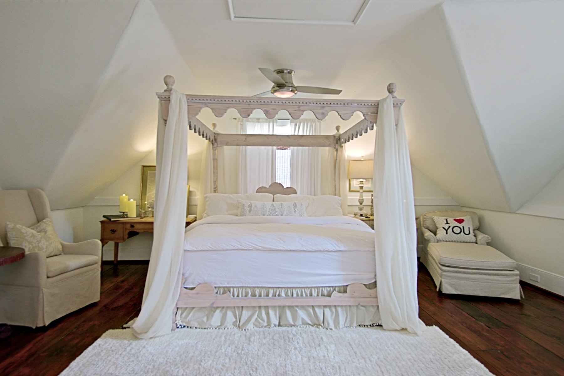 A symmetrical, wide-angle shot of an attic-style bedroom featuring a white four-poster canopy bed with flowing white curtains and a decorative scalloped frame. The room has sloped white ceilings and dark wood floors, with a beige armchair on the left and a matching chaise lounge on the right. A plush white rug sits at the foot of the bed, and a ceiling fan is visible above.