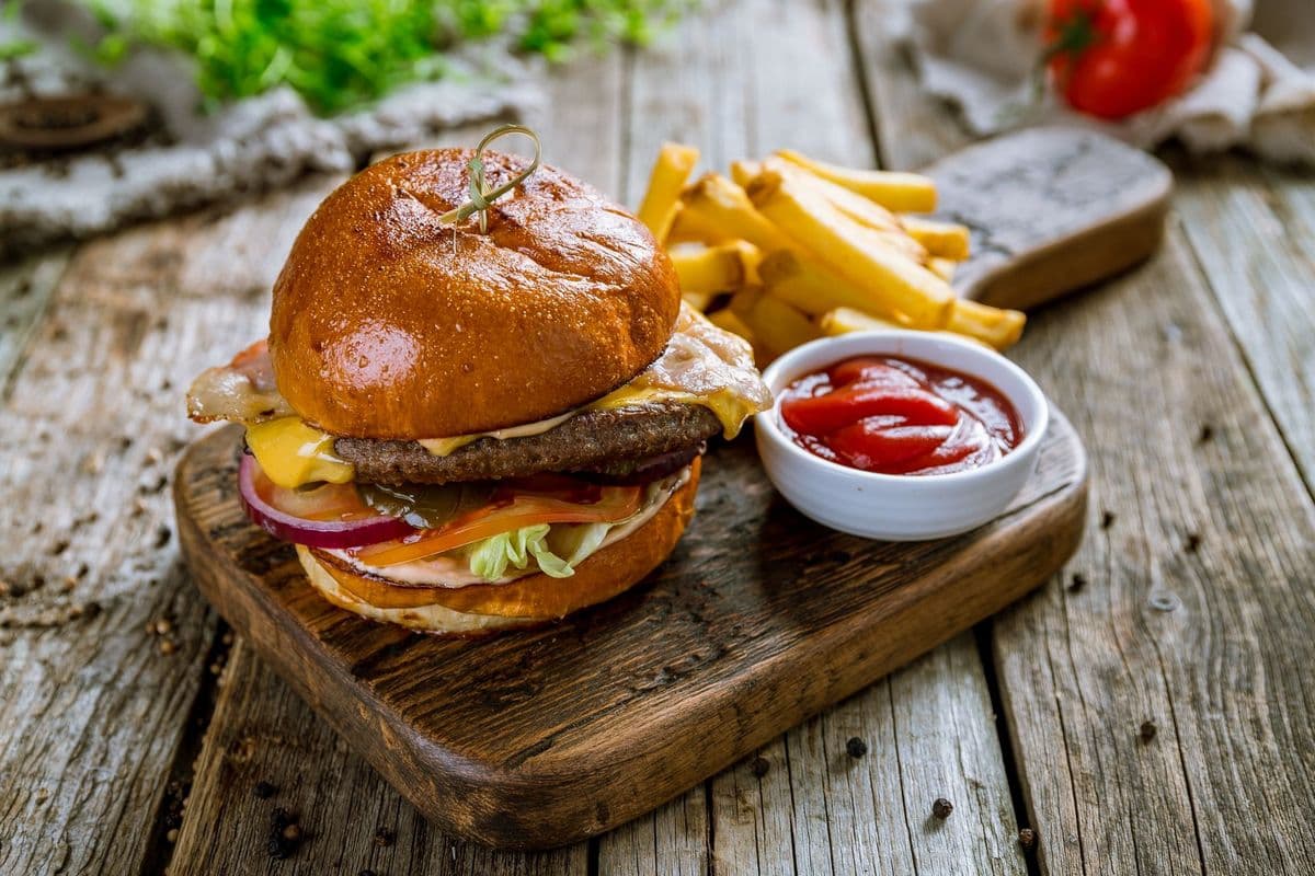A juicy burger loaded with cheese, lettuce, and onions, served with fries and ketchup on a wooden board.