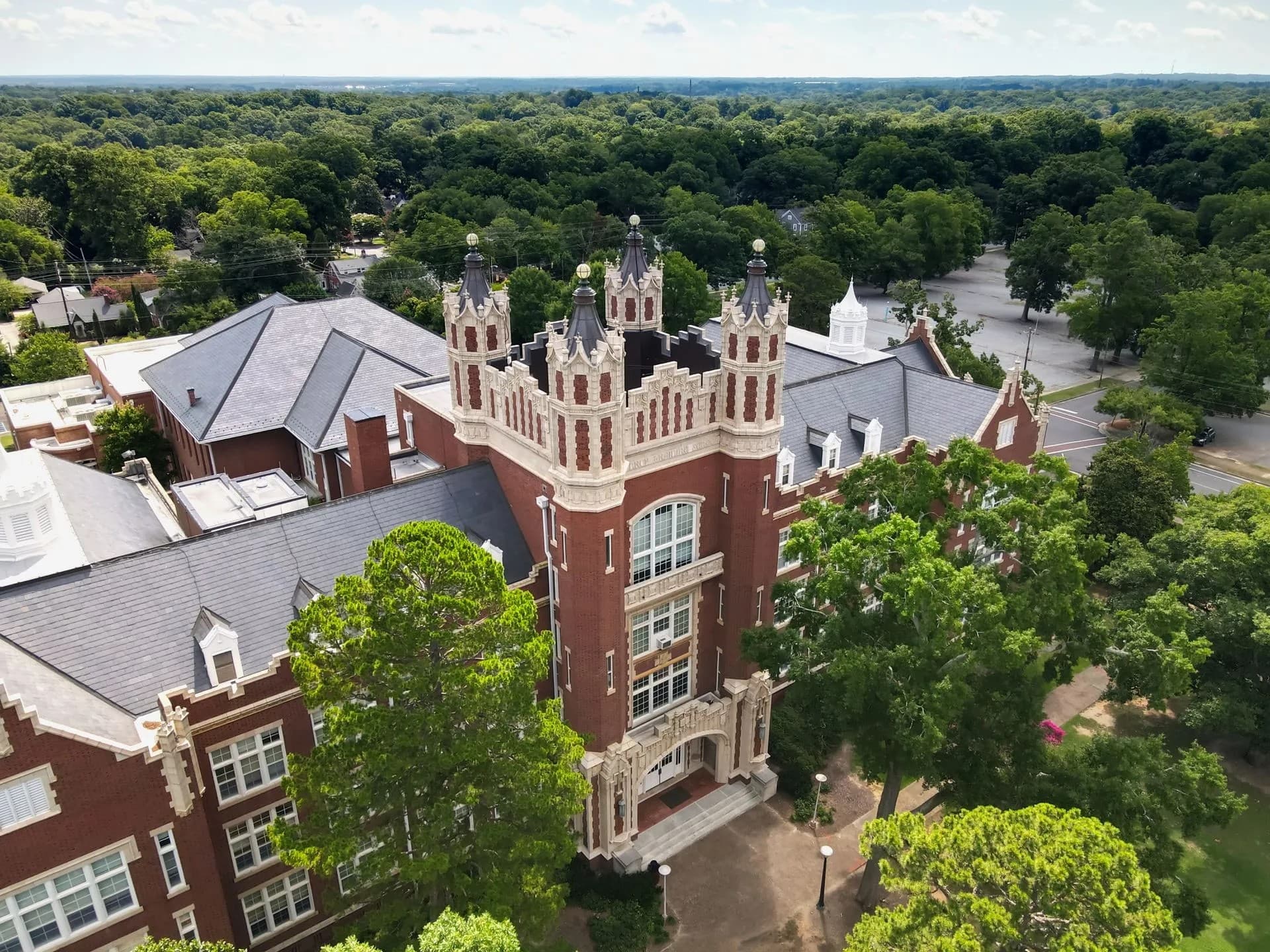 Aerial view of a historic red brick building with turret-like towers, surrounded by lush greenery. Aerial view of a historic red brick building with turret-like towers, surrounded by lush greenery.