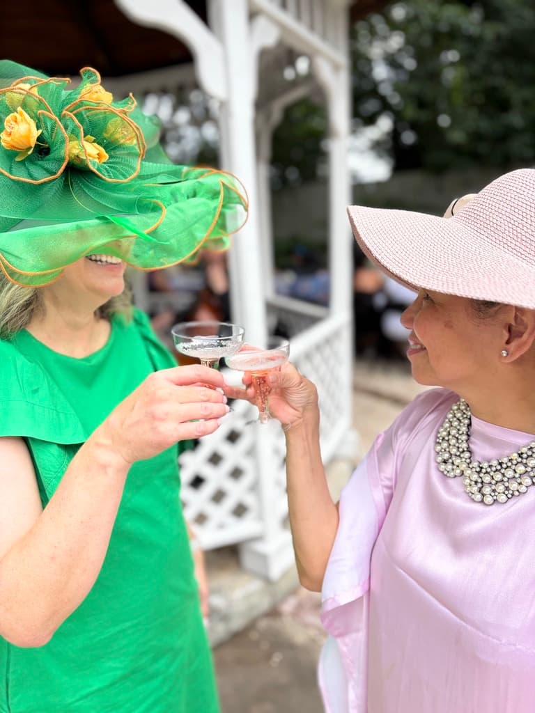 Two women clink champagne glasses while wearing stylish hats and smiling in a festive outdoor setting.