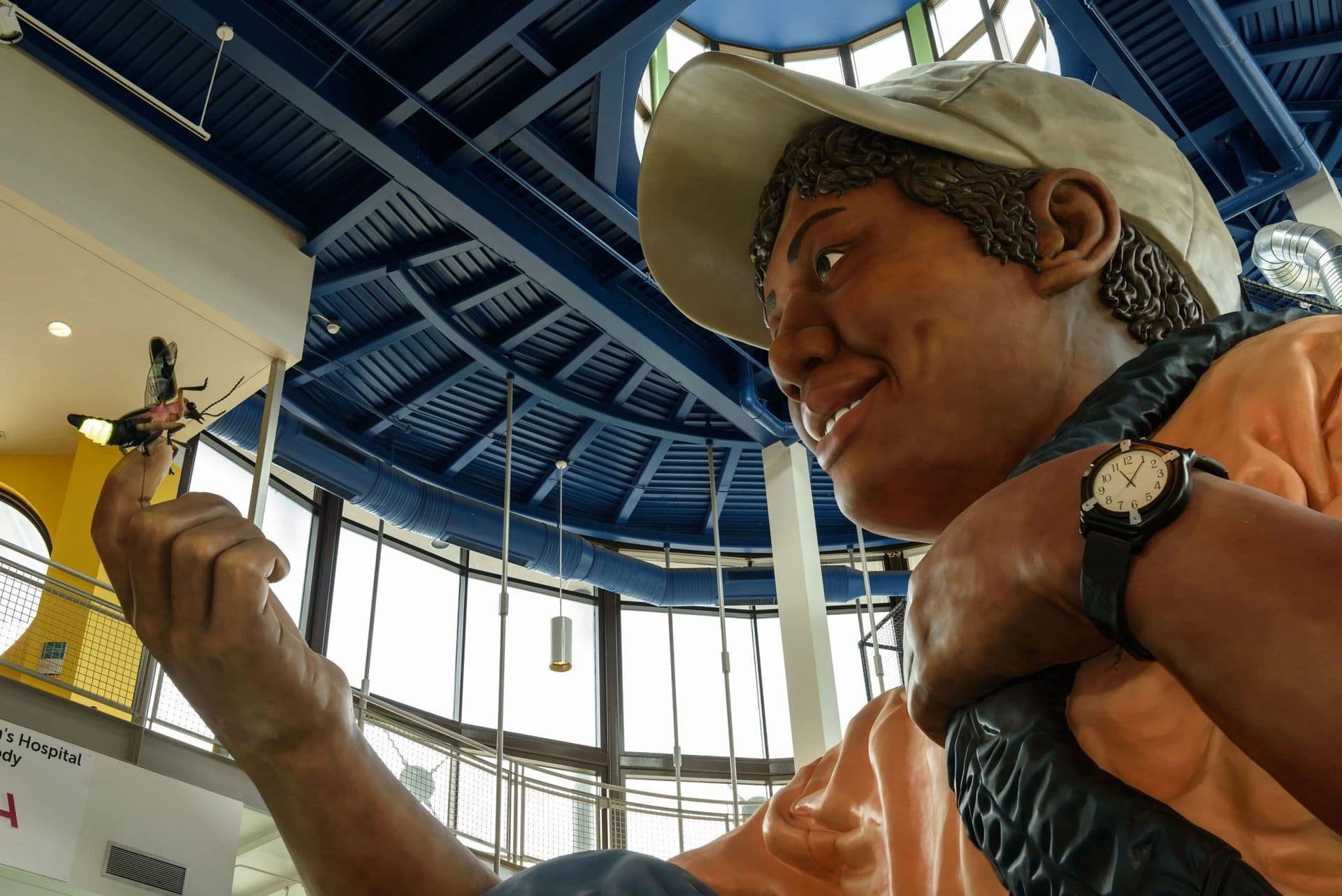 A large, smiling statue of a boy holding a model airplane, set in a brightly lit room with blue ceiling beams.