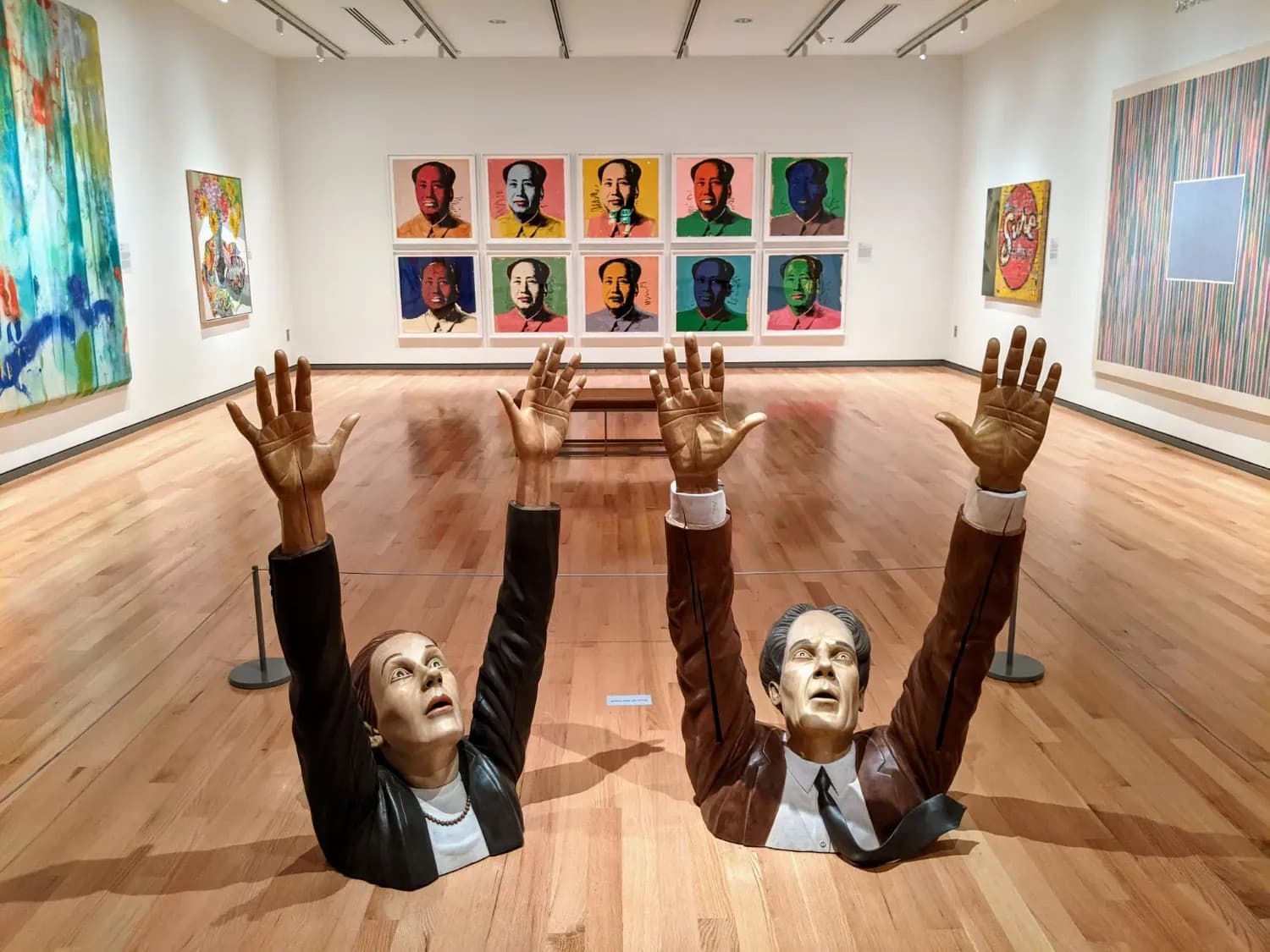 Two life-sized sculptures of a woman and man emerging from the floor with raised hands, set against colorful pop art portraits in a modern gallery.