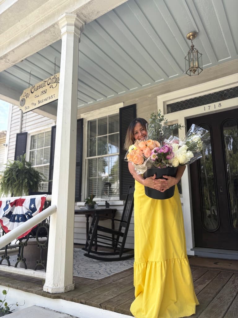 A woman in a yellow dress smiles while holding a large bouquet of flowers on the porch of a house.
