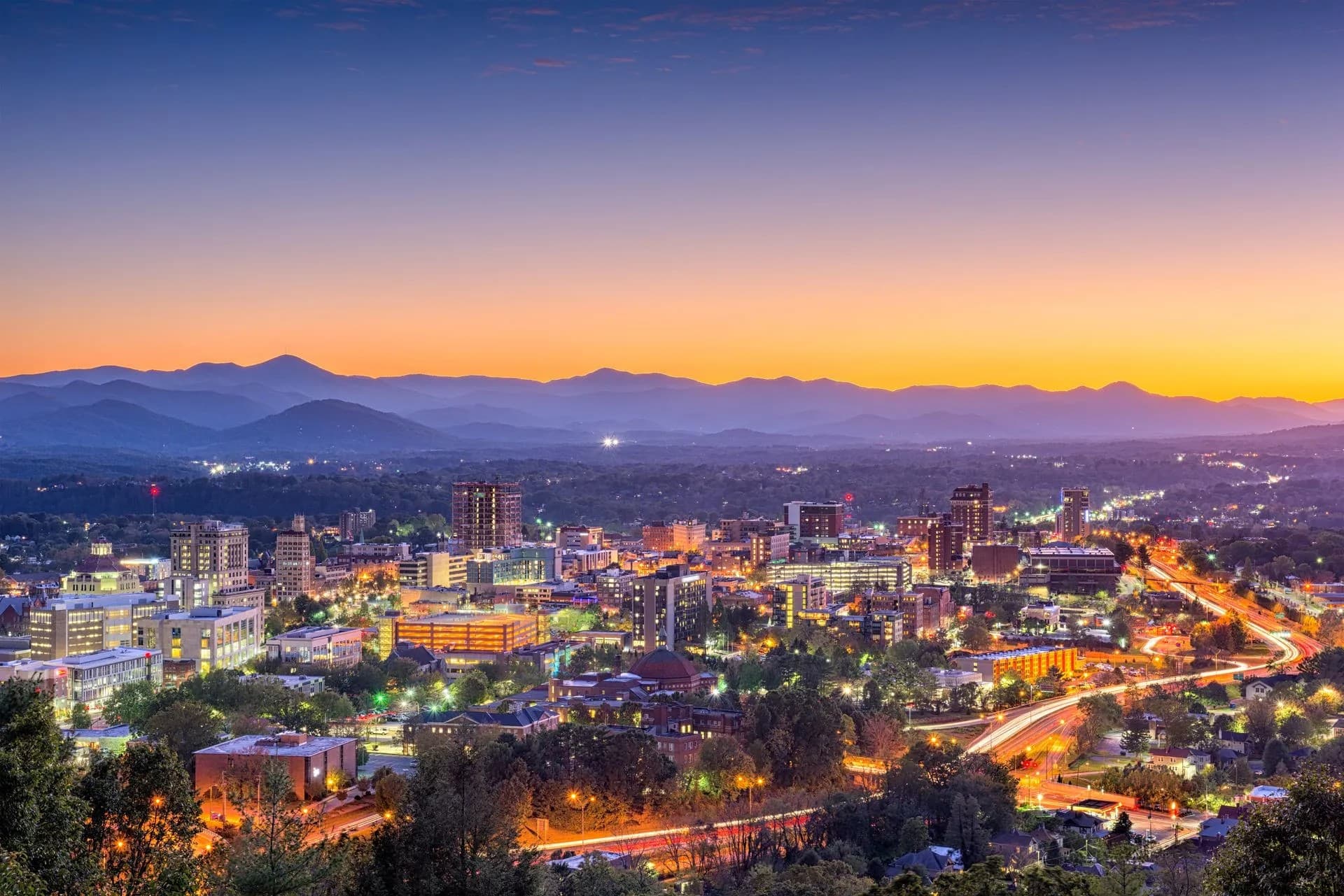 A panoramic view of a city skyline at dusk with mountains in the background and illuminated streets.