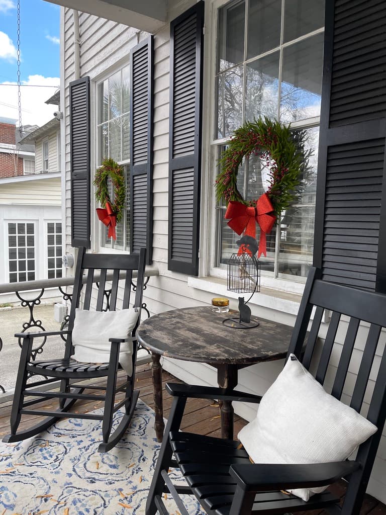 A cozy porch featuring two black rocking chairs, a rustic table, and festive wreaths adorned with red bows on the windows.