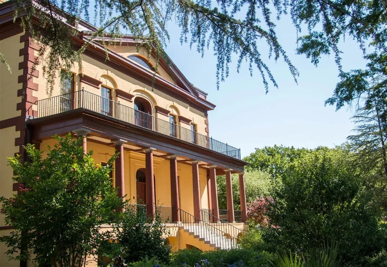 A grand, two-story yellow and red house with a wide staircase and balcony, surrounded by lush greenery.